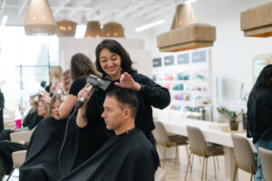 A stylist blow-dries a man's hair at the best salon in Waukee, with others enjoying balayage, highlights, and bridal hair services.