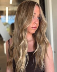 A woman with long, wavy, blonde balayage hair stands indoors, showcasing highlights and hair extensions from the best salon in Waukee.