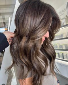 A client with long, wavy brunette hair, soft highlights, and loose layers at the best salon in Waukee for balayage and extensions.