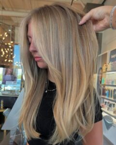 A woman with balayage blonde hair and highlights sits in the best salon in Waukee, as a stylist lifts her hair to show texture.