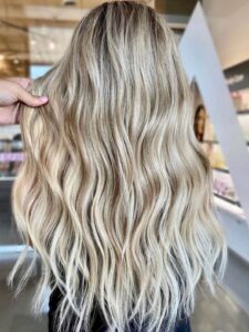 Inside the best salon in Waukee or Urbandale, balayage and highlights shine on long, wavy blonde hair; hair extensions shown.