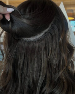 A stylist at the best salon in Waukee lifts brown hair to reveal balayage highlights and hidden hair extensions near the scalp.
