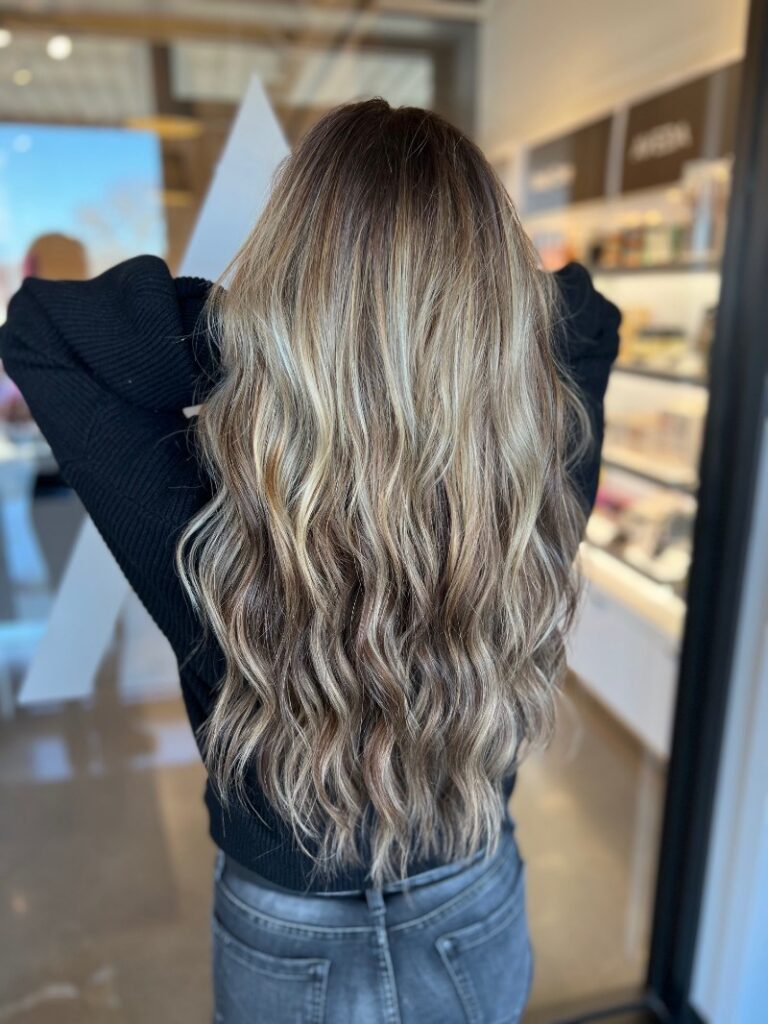 - Art of Life Salon & Spa at Urbandale and Waukee, IA A person with long, wavy balayage blonde hair and highlights stands indoors, showcasing best salon in Waukee and Urbandale style. - Art of Life Salon & Spa at Urbandale and Waukee, IA